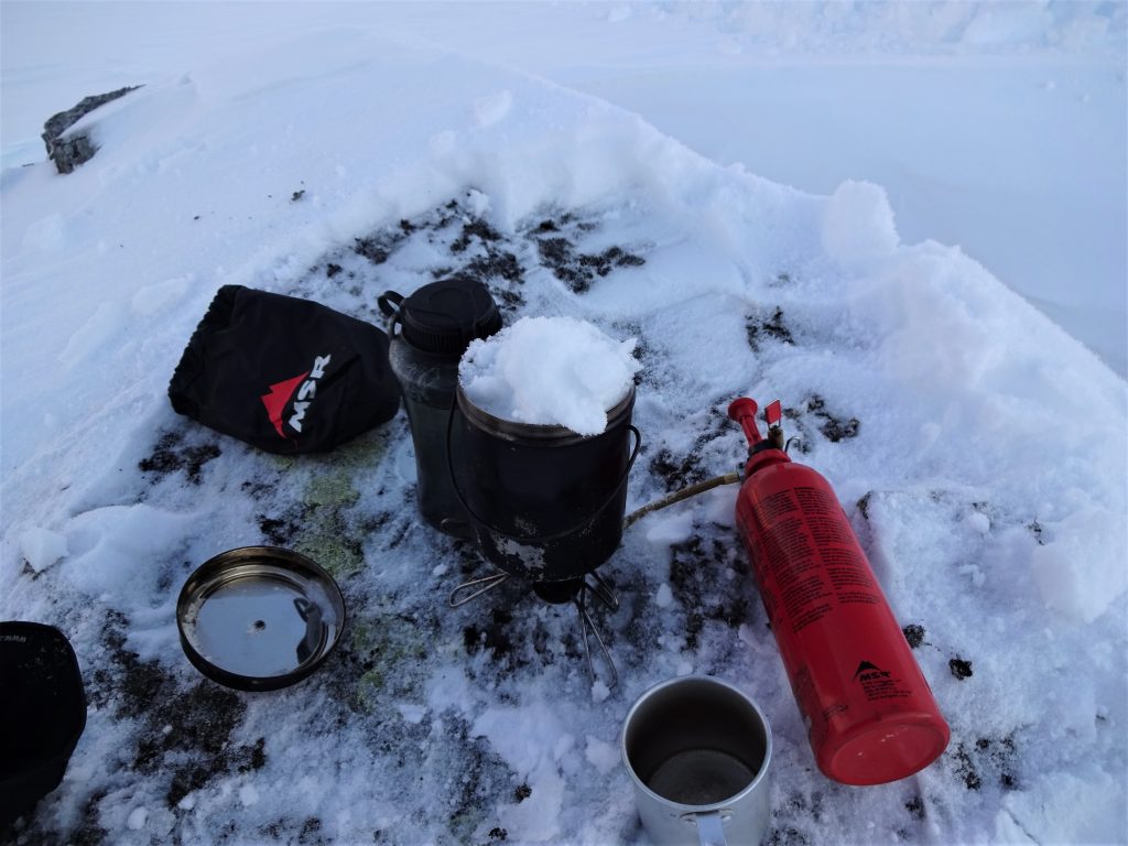 Op een rotssteen in een wit besneeuwd landschap staat een gasbrander met een billycan erop gevuld met sneeuw met daarnaast een aluminium beker.